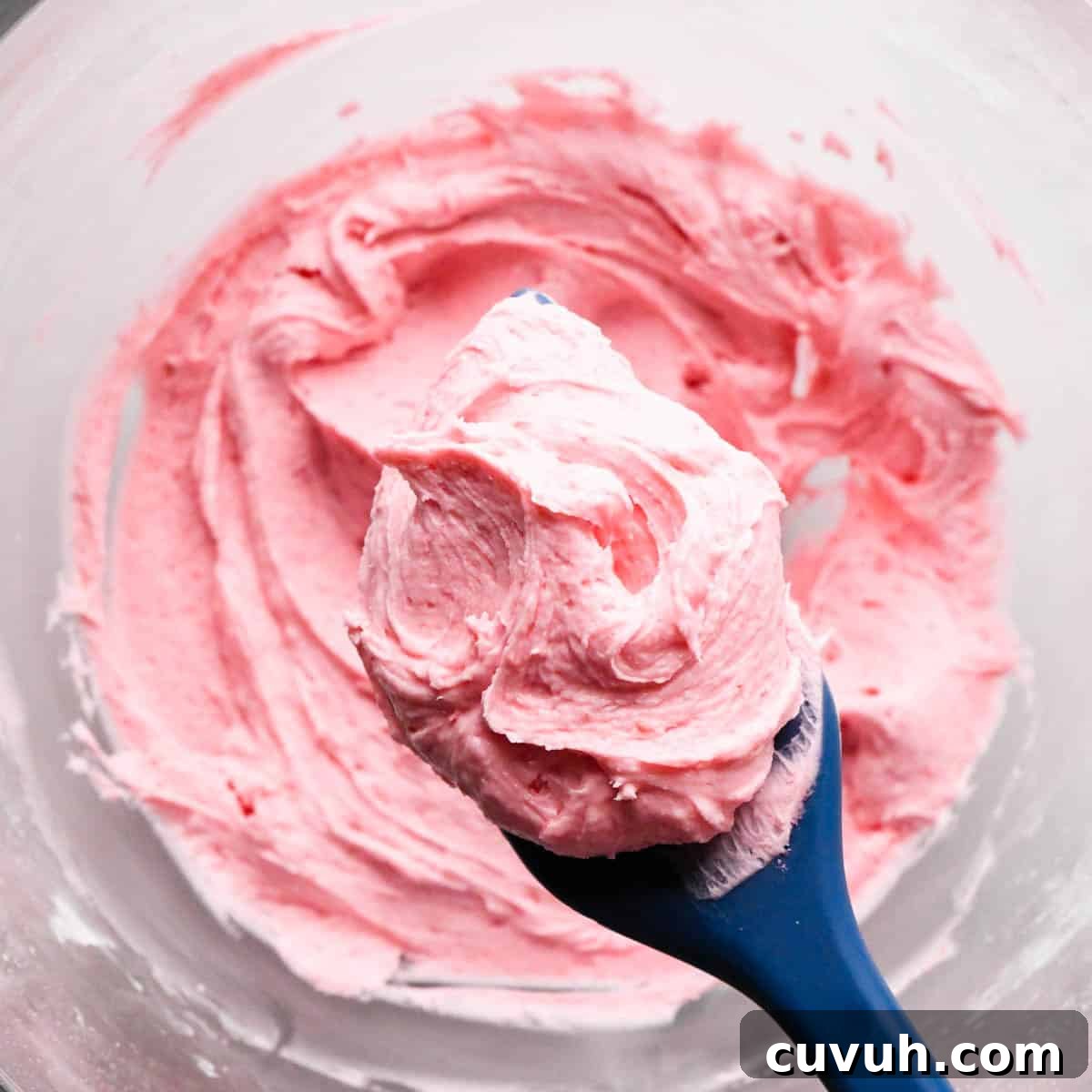 a blue spatula holding a scoop of raspberry frosting over a glass bowl of raspberry frosting