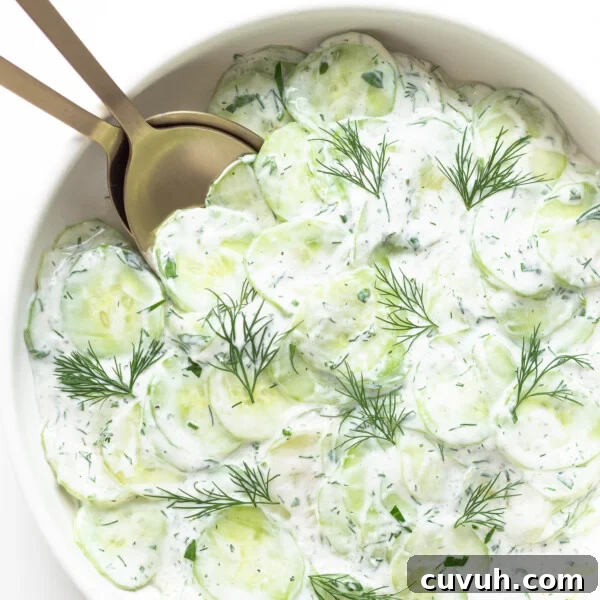 Close up of bowl containing creamy German cucumber salad (Gurkensalat) with gold salad servers.