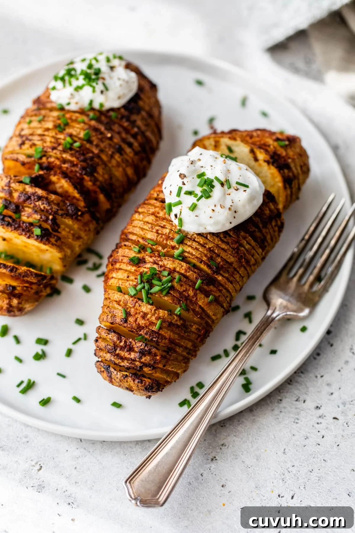 2 hasselback potatoes with sour cream and chives on top on a plate with a fork
