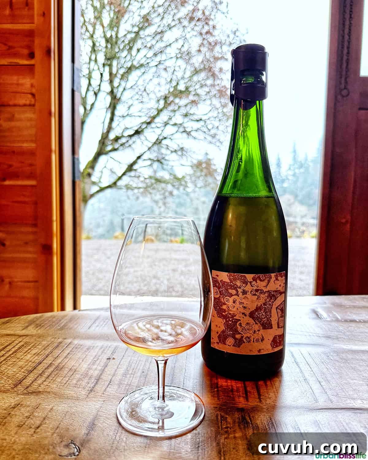 Bottle of Evening Land Vineyards Blanc de Noirs and glass of wine in front of cabin doors leading to the vineyard.