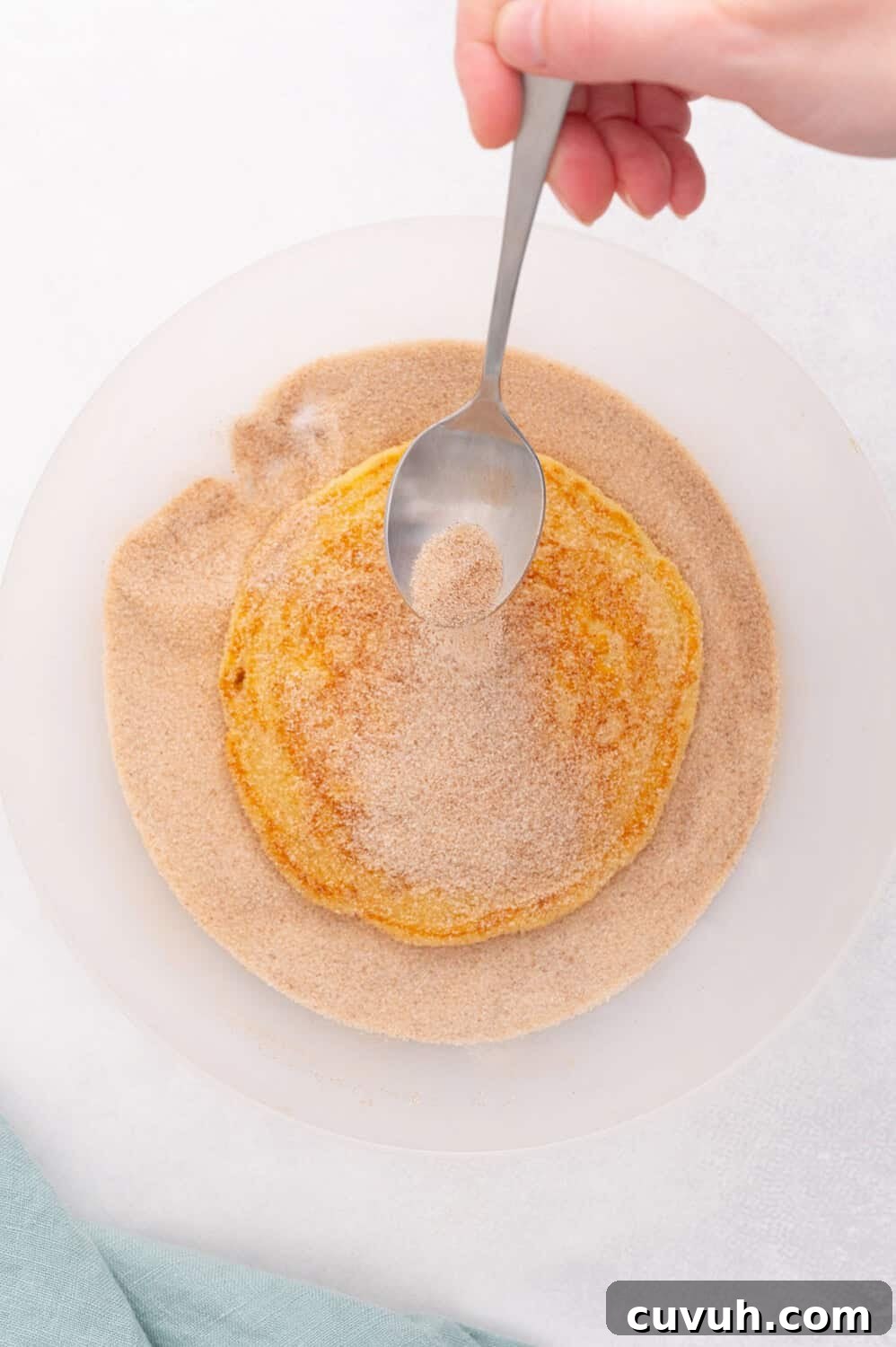 A hand holds a spoon, sprinkling sugar and cinnamon over a piece of bread in a white bowl on a white surface.