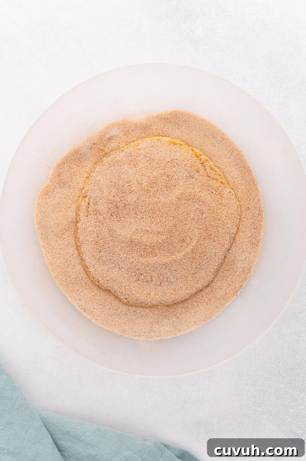 A pancake covered in cinnamon sugar rests on a plate, with a green cloth partially visible in the bottom left corner.