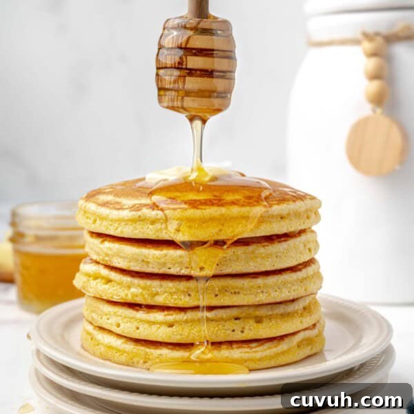 A stack of pancakes on a plate with butter on top, being drizzled with honey.