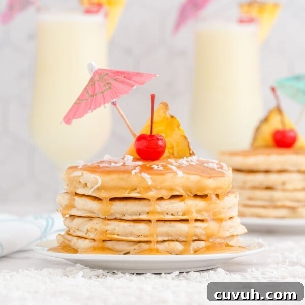 A stack of pancakes topped with caramel sauce, a pineapple slice, a cherry, shredded coconut, and a paper drink umbrella.