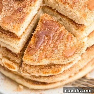 A stack of fluffy churro pancakes, drizzled with syrup, and sprinkled with cinnamon sugar, with a triangle slice pulled out, on a white plate.