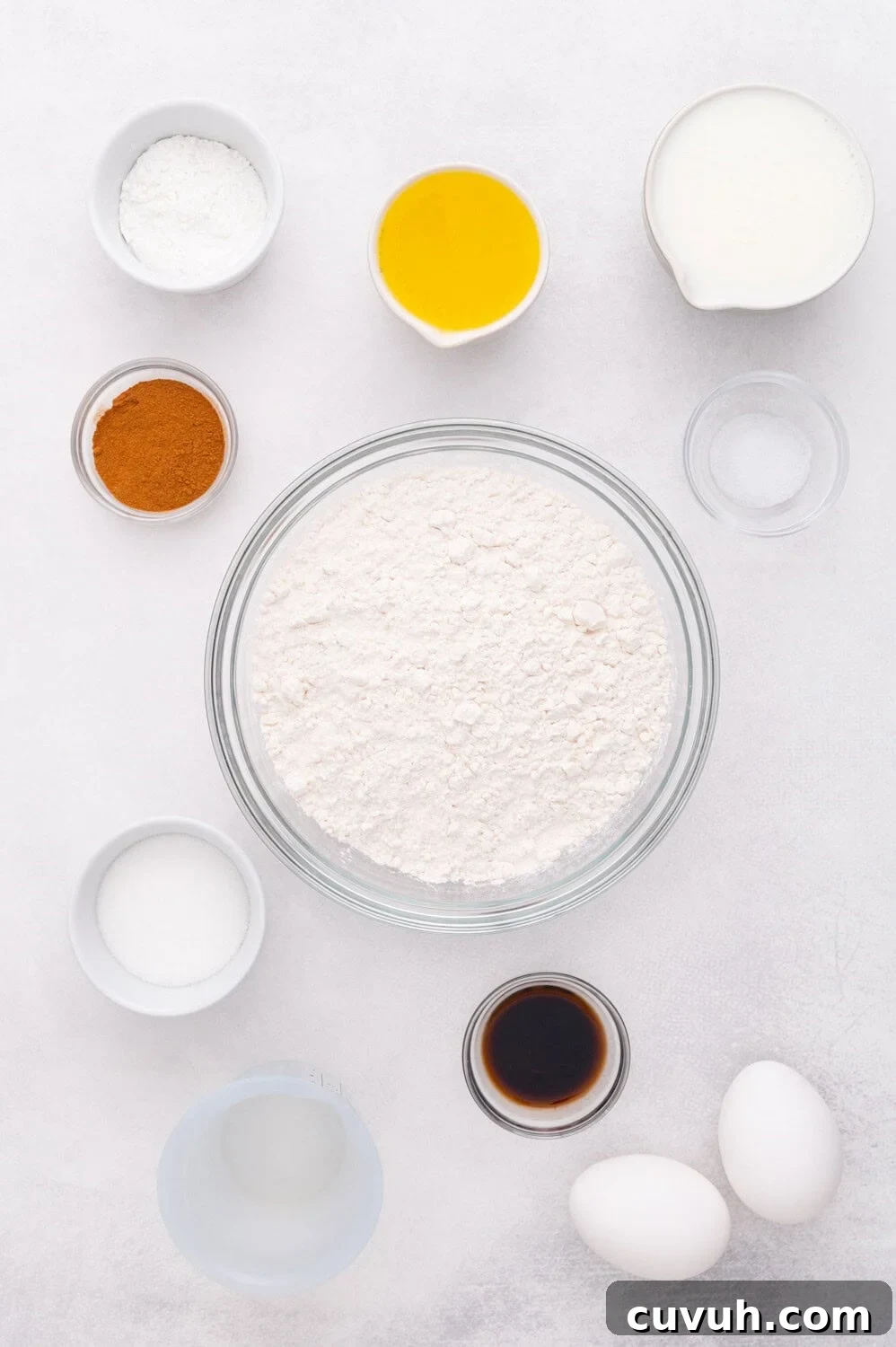 A variety of ingredients for baking are arranged on a white surface, including flour, eggs, vanilla, oil, sugar, cinnamon, baking powder, and milk.