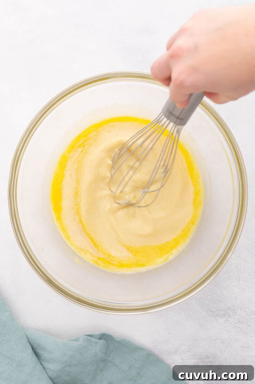 Hand whisking yellow batter in a clear glass bowl on a white surface, with a blue cloth nearby.