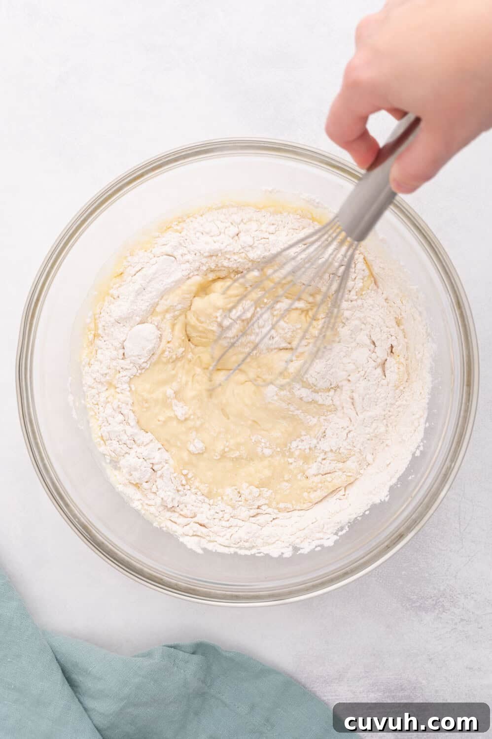 A hand whisking a mixture of flour and liquid in a clear glass bowl, placed on a light-colored surface with a green cloth nearby.