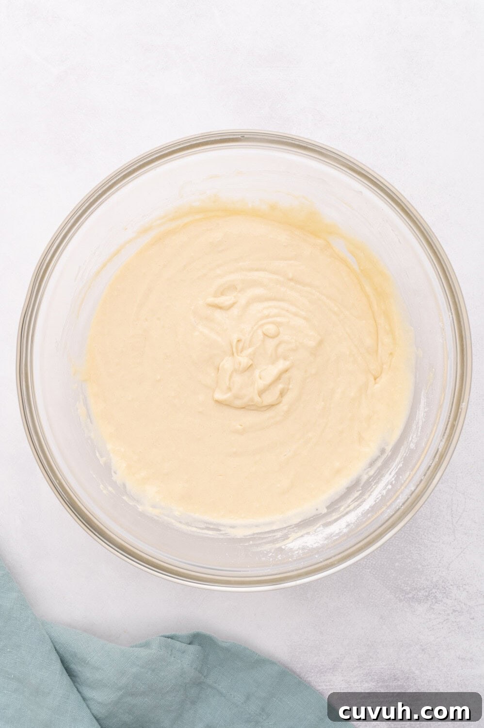 A glass bowl with smooth, creamy pancake batter sits on a light surface next to a folded blue cloth.
