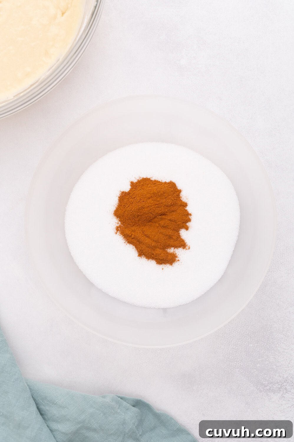 A bowl containing granulated sugar and a mound of cinnamon, next to a green cloth and a mixing bowl.