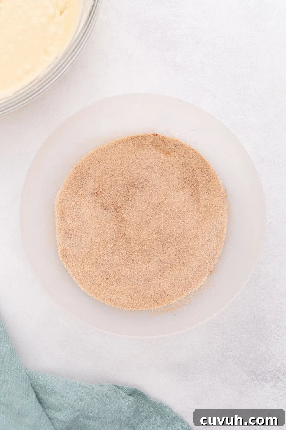 A bowl filled with cinnamon sugar mixture on a light surface. A corner of a blue cloth is visible next to the bowl.