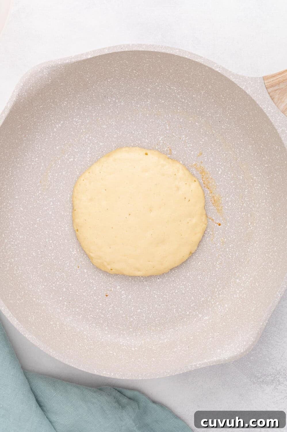 A single pancake cooking in a non-stick frying pan, with a light tan surface. A folded green kitchen towel is partially visible at the side.