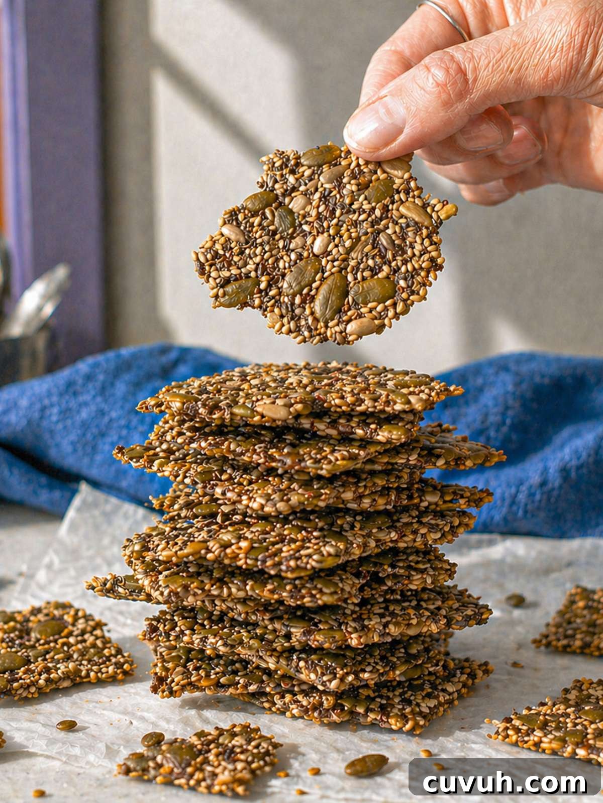 Crispy Seed Crackers: Flour-Free Savory Snack Recipe 2 Hand holding a thin seed cracker above a tall stack of crispy seed crackers in natural light.