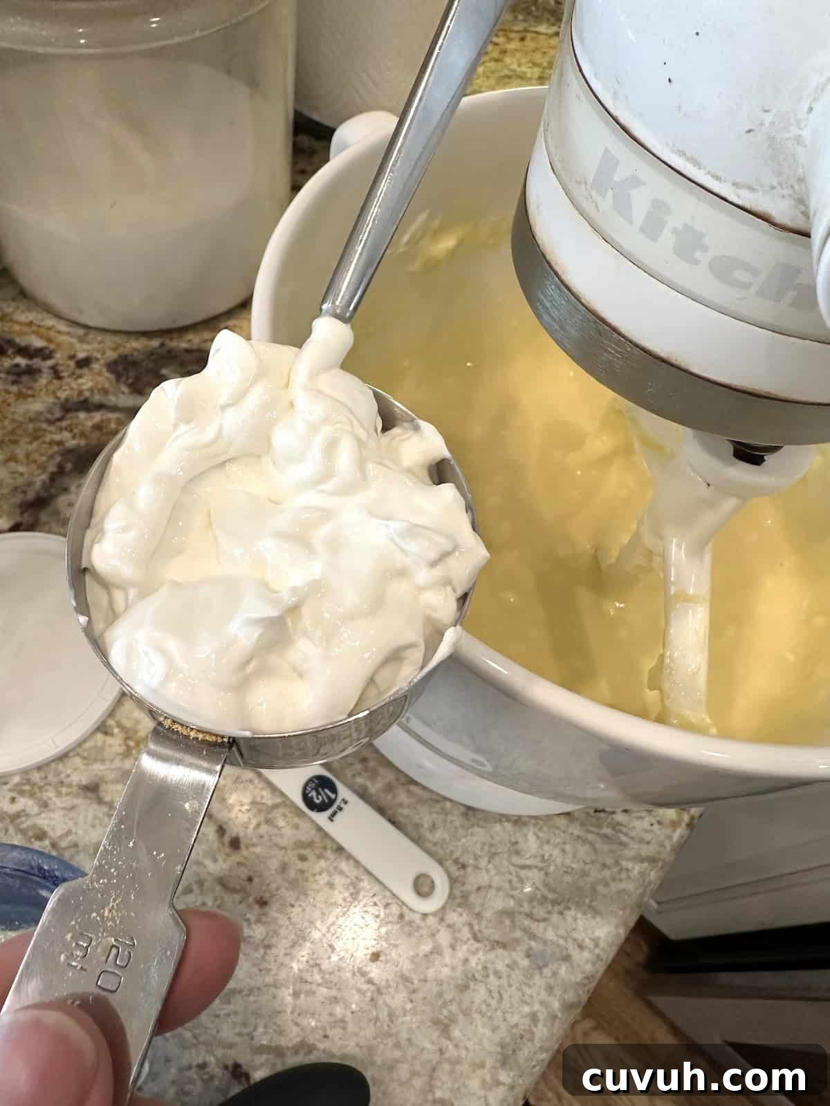 A measuring scoop with sour cream near a mixing bowl with cream cheese filling ingredients for cheesecake.