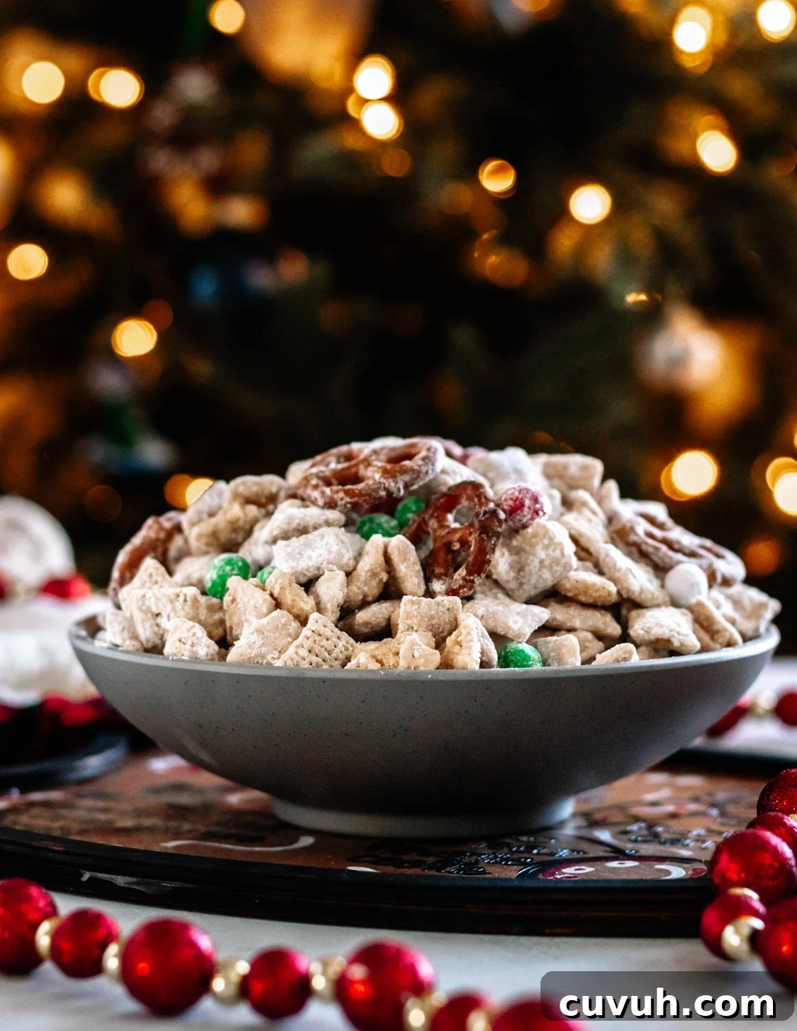 Christmas Puppy Chow Recipe — Festive Reindeer Munch Treats 2 Give Santa what he wants! A bowl of Christmas Muddy Buddy Mix in a grey bowl set in front of a lit Christmas tree