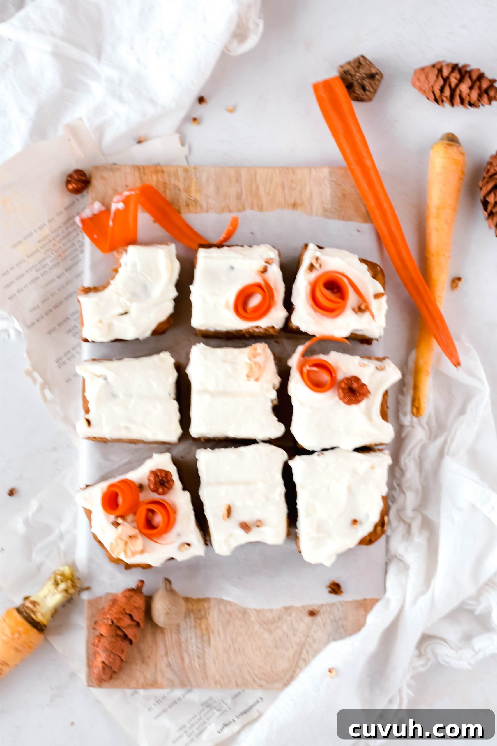 Overhead view of 9 square carrot cake bars topped with cream cheese icing and shredded carrots on a cutting board lined with parchment paper