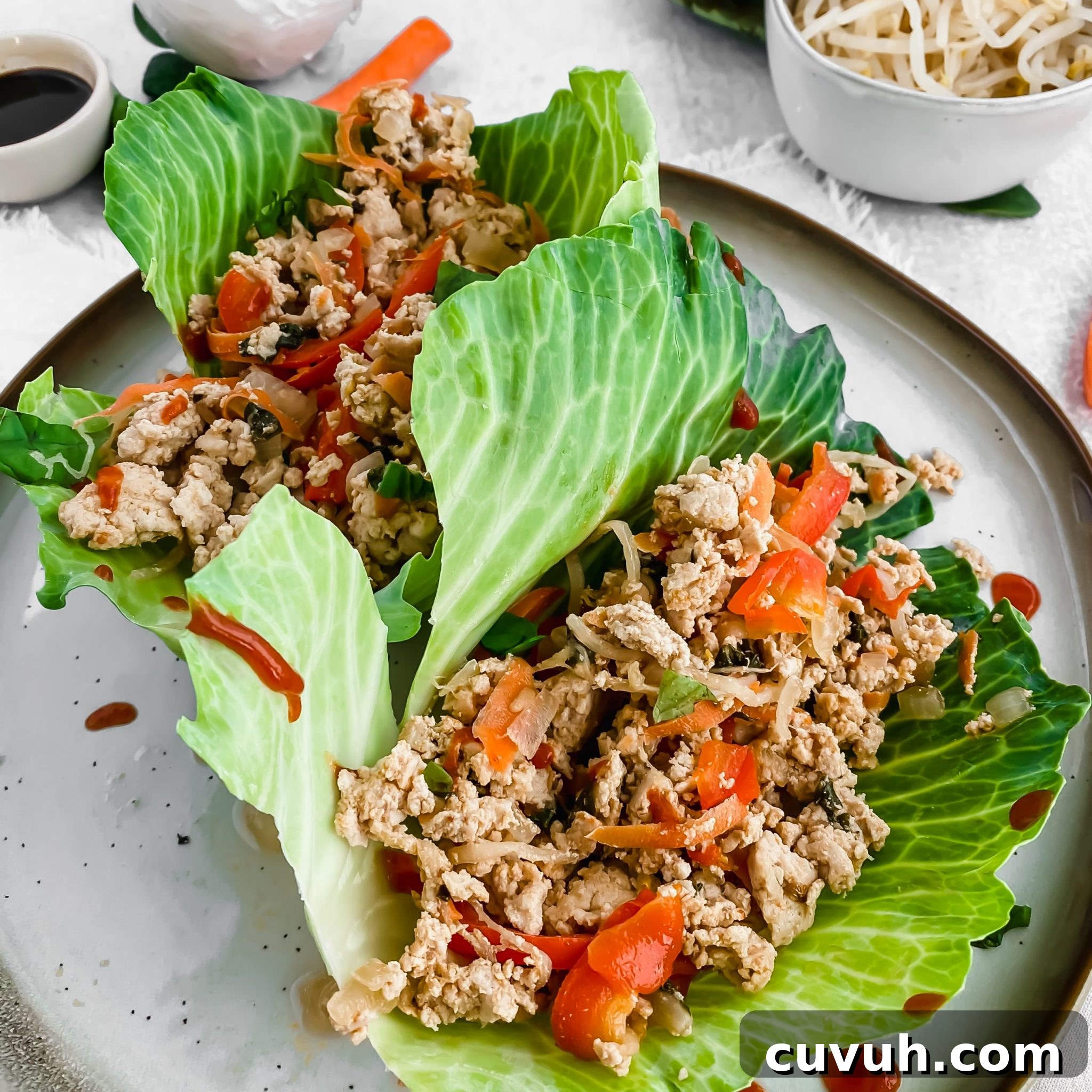 two Thai chicken lettuce wraps sitting on a ceramic plate with sriracha drizzled overtop.