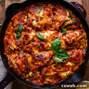 A cast iron pan with eggplant lasagna baked in it. The pan is placed in a wooden background with fresh herbs and a parmesan rind arranged around it.
