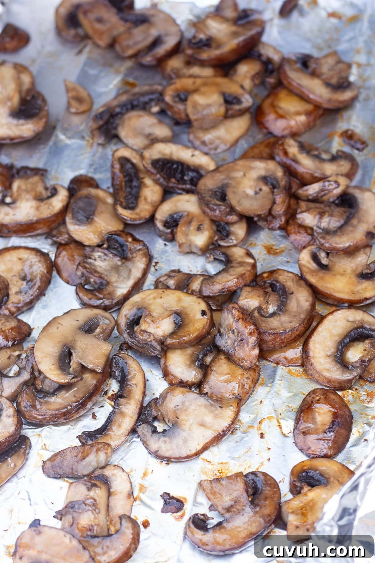 Grilled Foil-Wrapped Mushrooms with Garlic and Herbs 11 A close-up of grilled mushroom slices on a piece of aluminum foil that's resting on grill grates. The mushrooms appear to be cooked, displaying a browned and slightly crisp exterior. The aluminum foil underneath is lightly stained with cooking juices and oil.