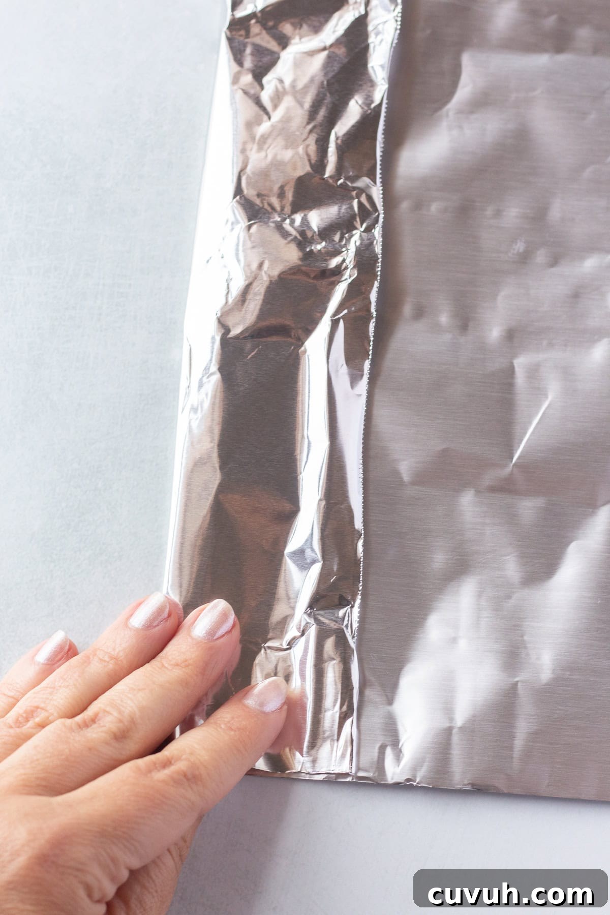 Grilled Foil-Wrapped Mushrooms with Garlic and Herbs 5 A hand with light-colored nails is shown folding the edge of a sheet of aluminum foil on a light surface. The foil has a shiny, reflective texture and a slightly crumpled appearance near the fold.