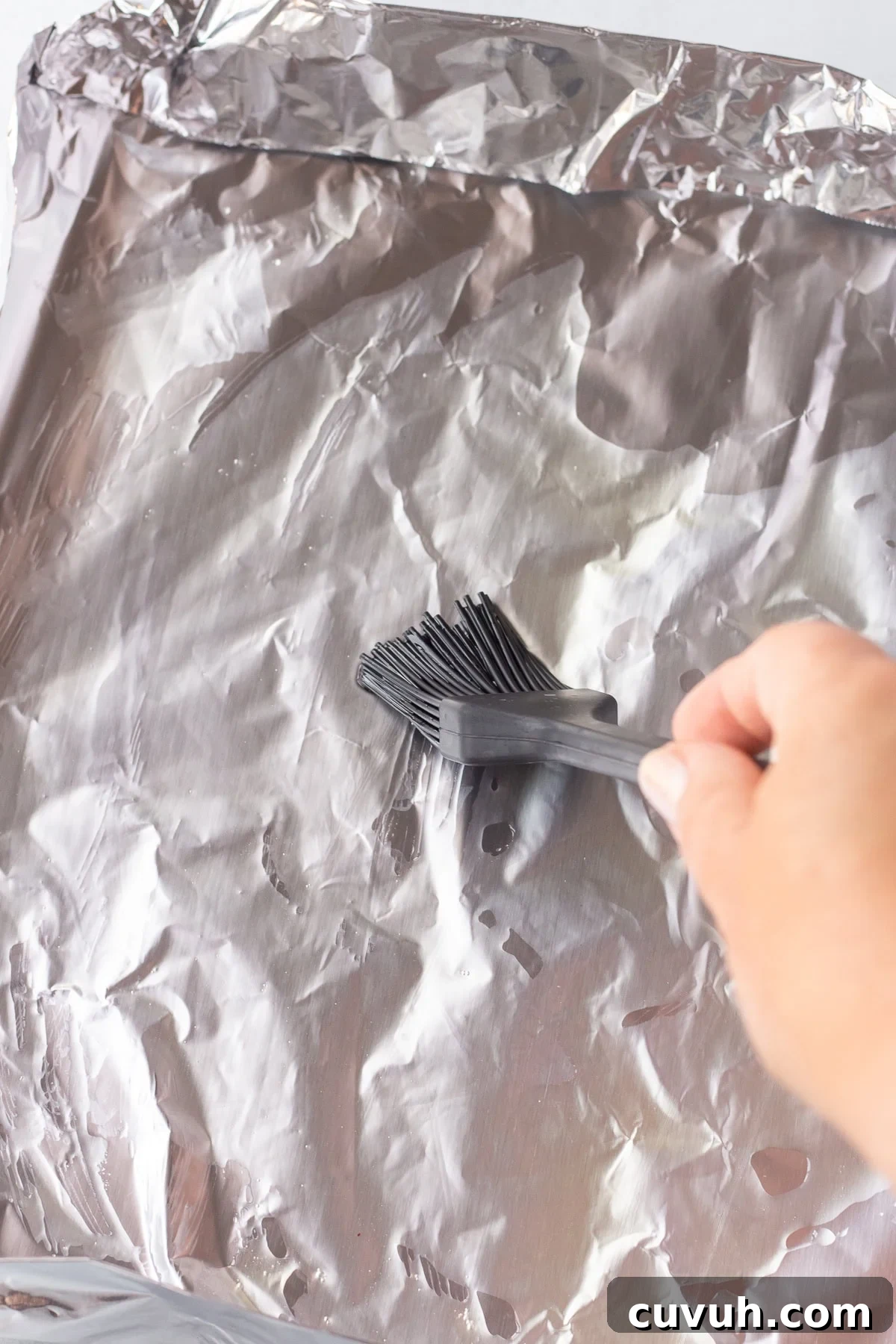 Grilled Foil-Wrapped Mushrooms with Garlic and Herbs 7 A hand holds a brush to spread oil or another substance over a sheet of aluminum foil folded into a baking tray. The foil is crinkled and reflects light. The brush has black bristles and a grey handle.