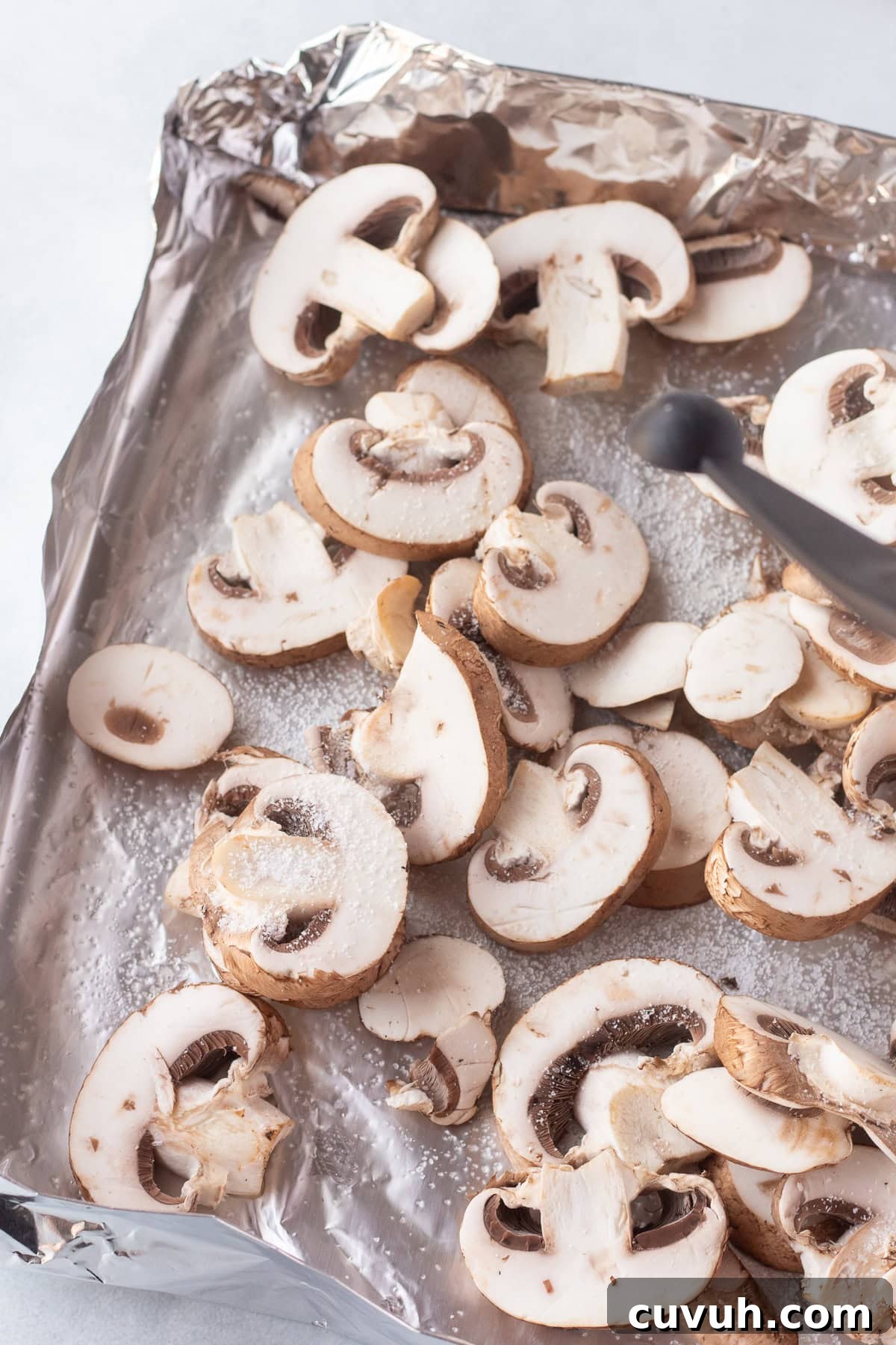 Grilled Foil-Wrapped Mushrooms with Garlic and Herbs 8 A piece of aluminum foil made into a pan is covered with sliced mushrooms. The mushrooms appear to be freshly cut and slightly seasoned. A pair of tongs is visible on the side of the baking sheet.