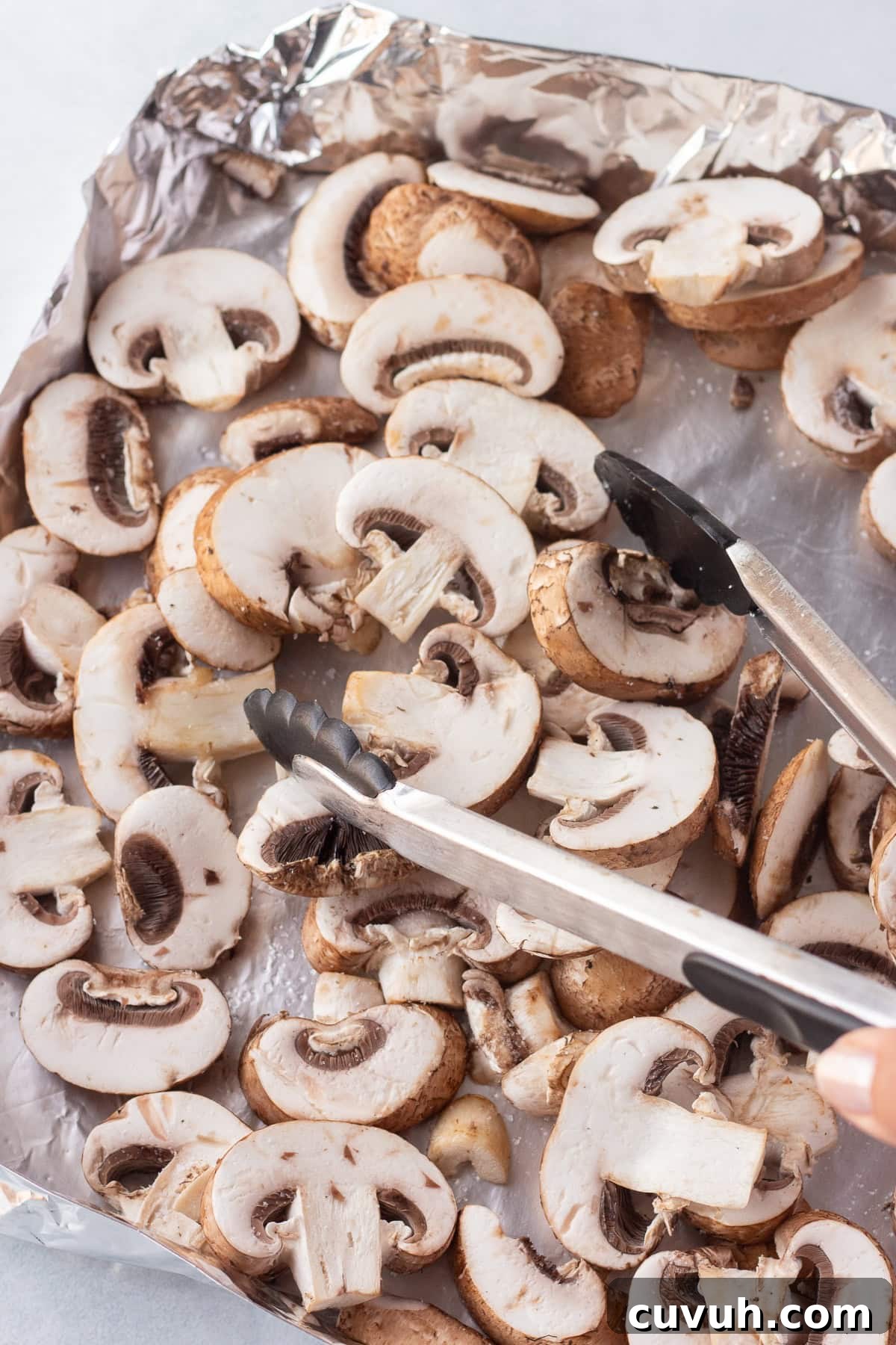 Grilled Foil-Wrapped Mushrooms with Garlic and Herbs 9 A piece of aluminum foil folded into a pan shape holds sliced mushrooms. A pair of tongs on the right side of the image is being used to arrange the mushrooms. The background is neutral and out of focus.