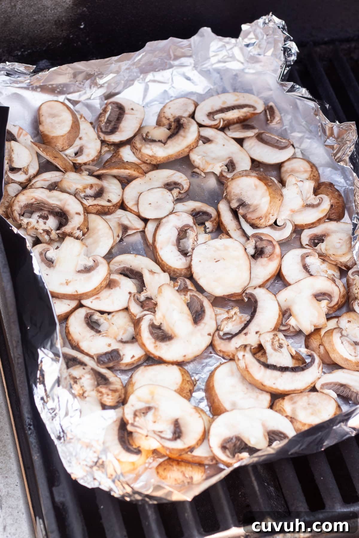 Grilled Foil-Wrapped Mushrooms with Garlic and Herbs 10 A sheet of aluminum foil on a grill is topped with sliced mushrooms. The mushrooms appear to be slightly cooked, lying evenly spread across the foil.