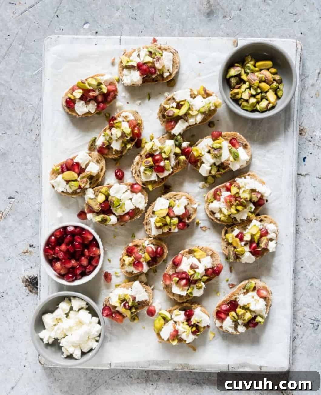 Thanksgiving Appetizers You Can Make Ahead for Stress-Free Hosting 8 Crostinis with pomegrant, cheese and pistachios on a white serving board.