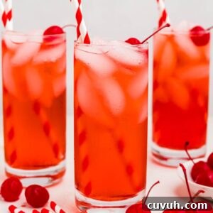 Bright red drink in a tall glass with ice and cherries on the table.