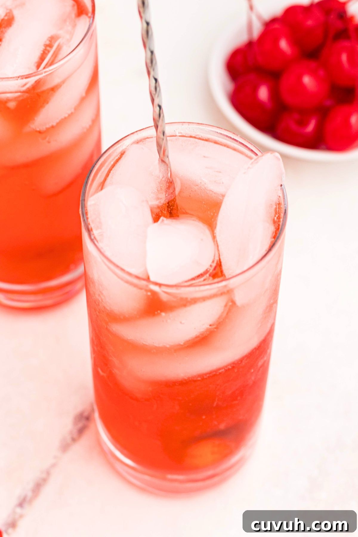 Bright red drink in a tall glass with ice and cherries on the table.