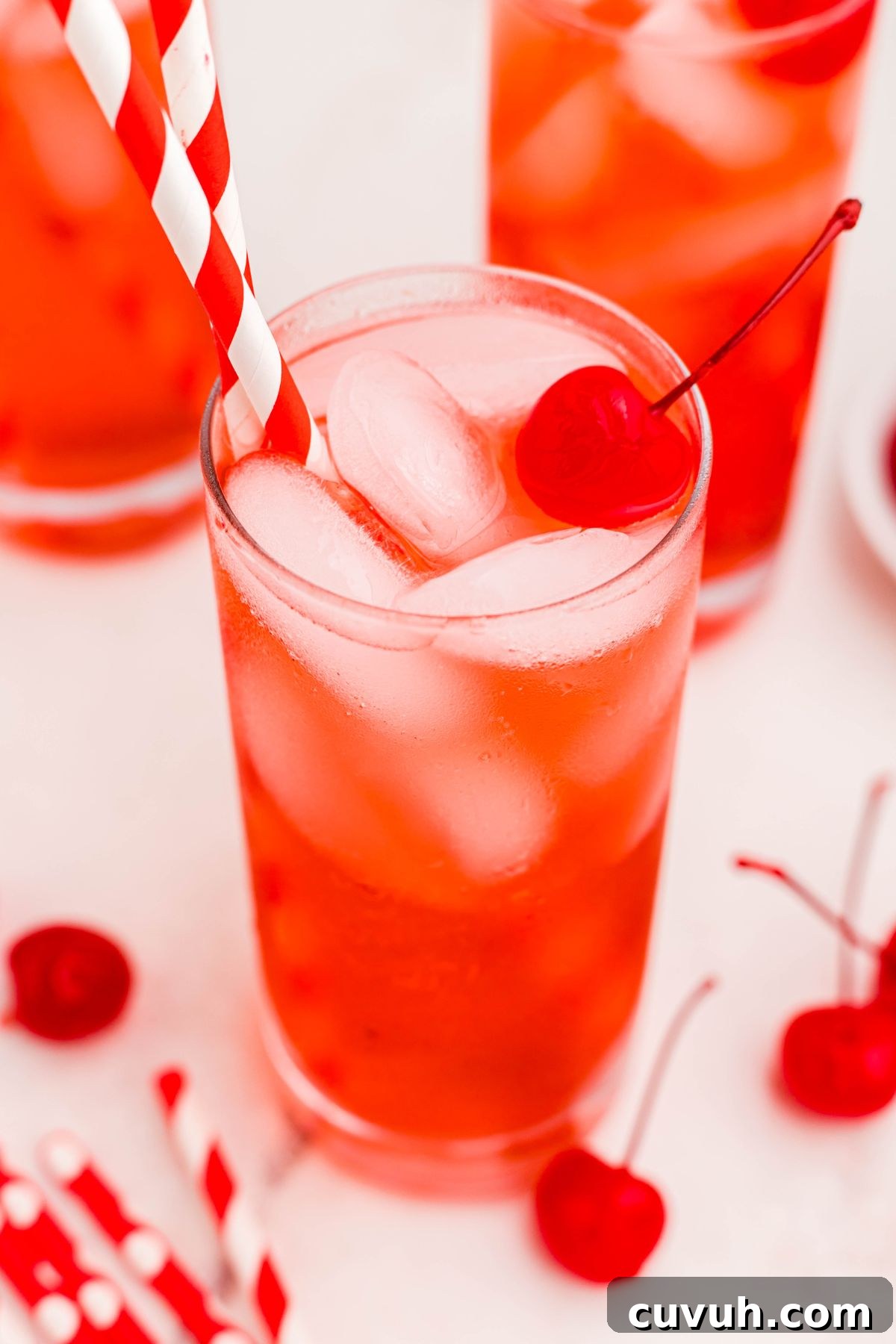 Bright red drink in a tall glass with ice, straws, and a cherry on top, and cherries on the table.