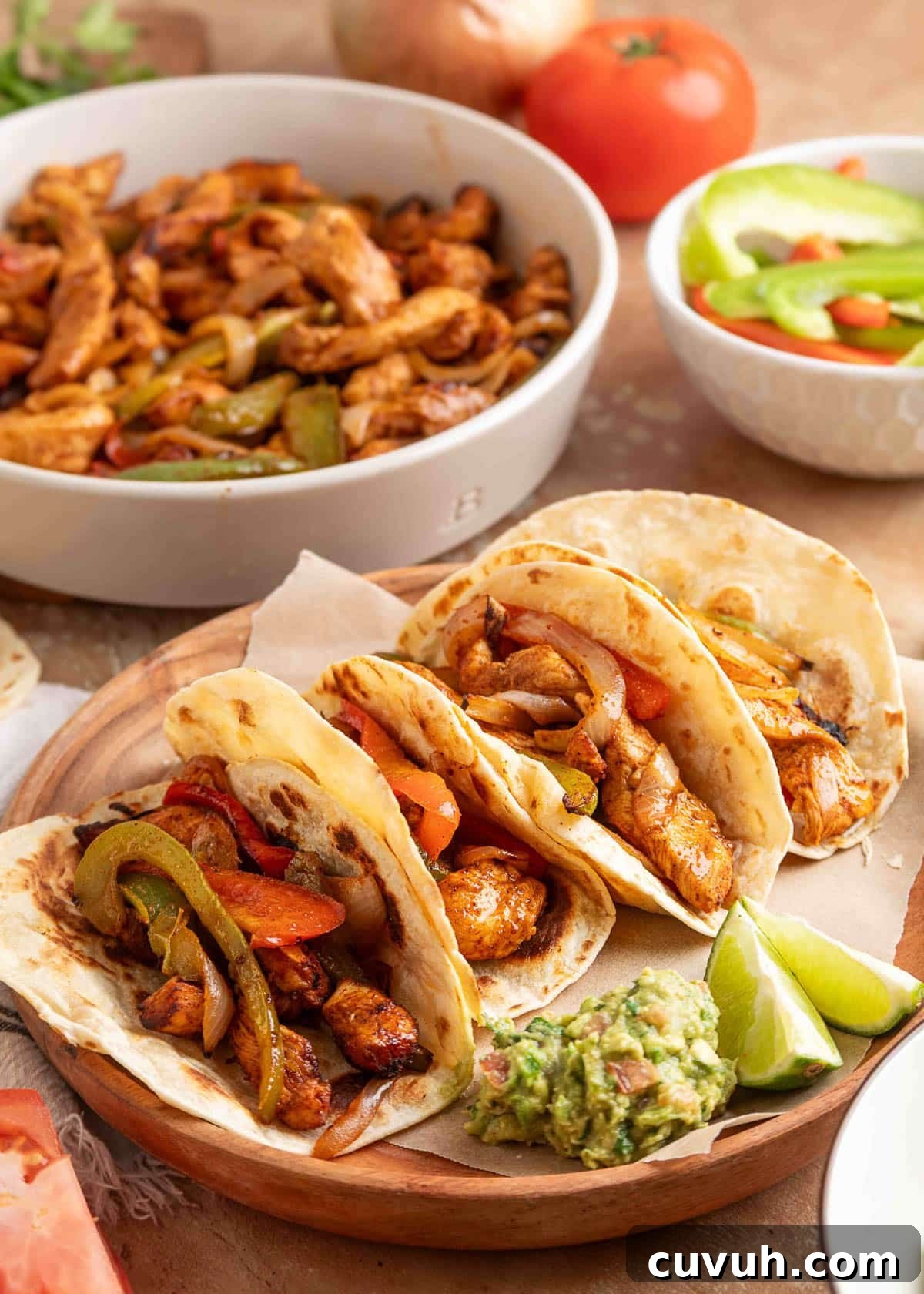 Crispy Air Fryer Chicken Fajitas with Peppers and Onions 2 Four flour tortillas filled with air fryer chicken fajitas, including seasoned chicken strips, sautéed onions, and bell peppers, served on a wooden plate with lime wedges and a scoop of chunky guacamole. A bowl of extra fajita filling and sliced peppers are visible in the background.
