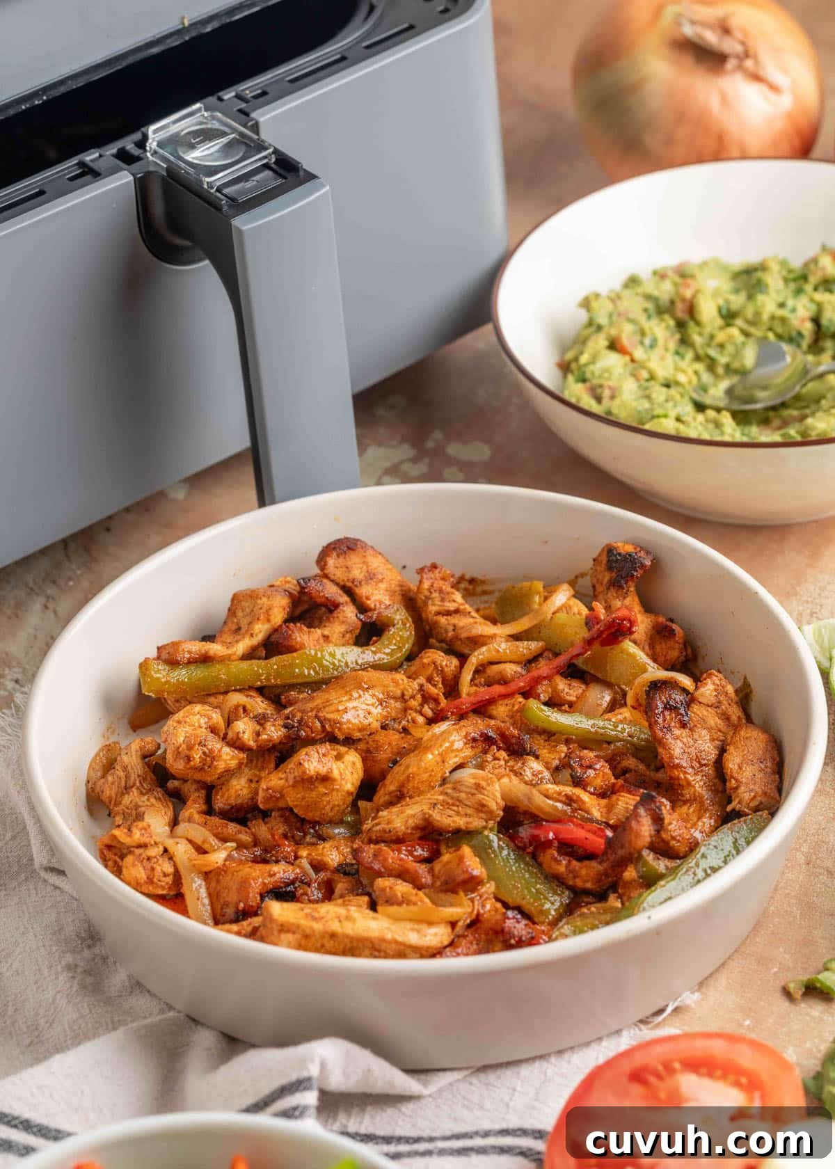 Crispy Air Fryer Chicken Fajitas with Peppers and Onions 12 Bowl of air fryer chicken fajitas with sautéed peppers and onions, set on a cloth next to an air fryer and a bowl of guacamole.