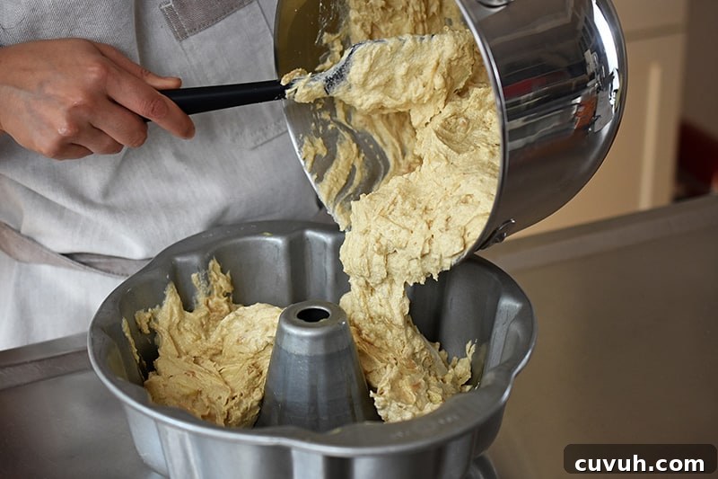 Someone using a black silicone spatula to transfer the apple Bundt Cake batter into a greased bundt pan.