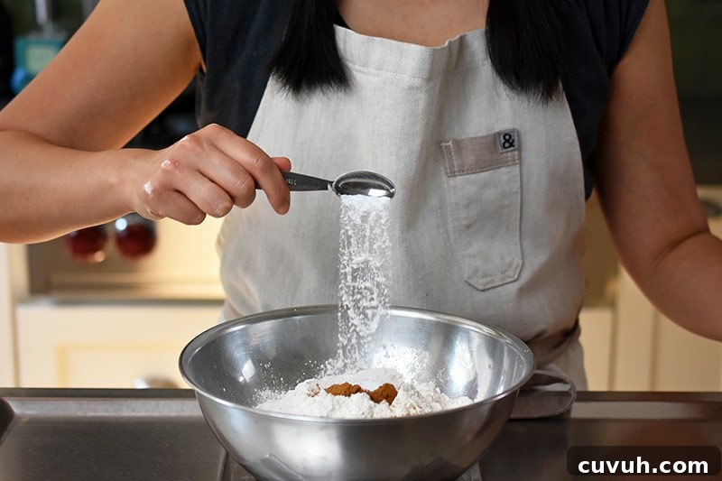 Someone adding a spoonful of baking soda to a bowl filled with the dry ingredients for apple Bundt Cake.