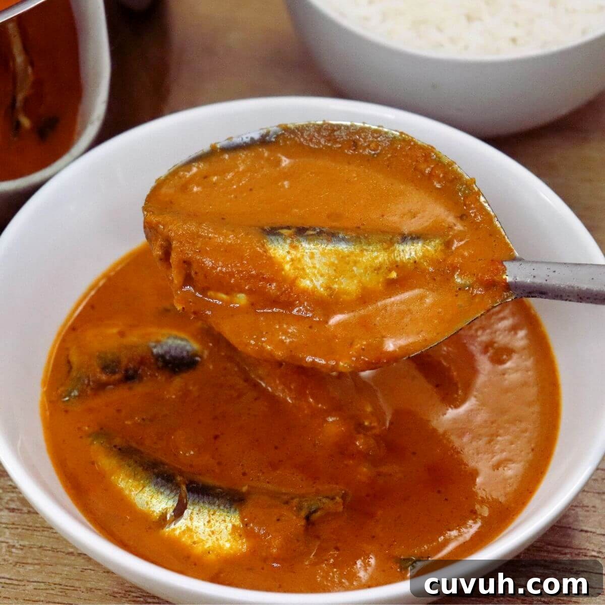 a ladle full of sardine curry is held over a bowl of sardine curry.