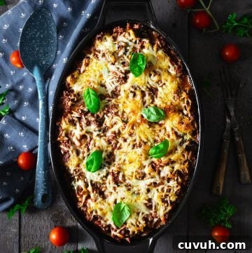 Ravioli lasagna in a black casserole dish on a gray wooden background with blue towel and tomatoes around.