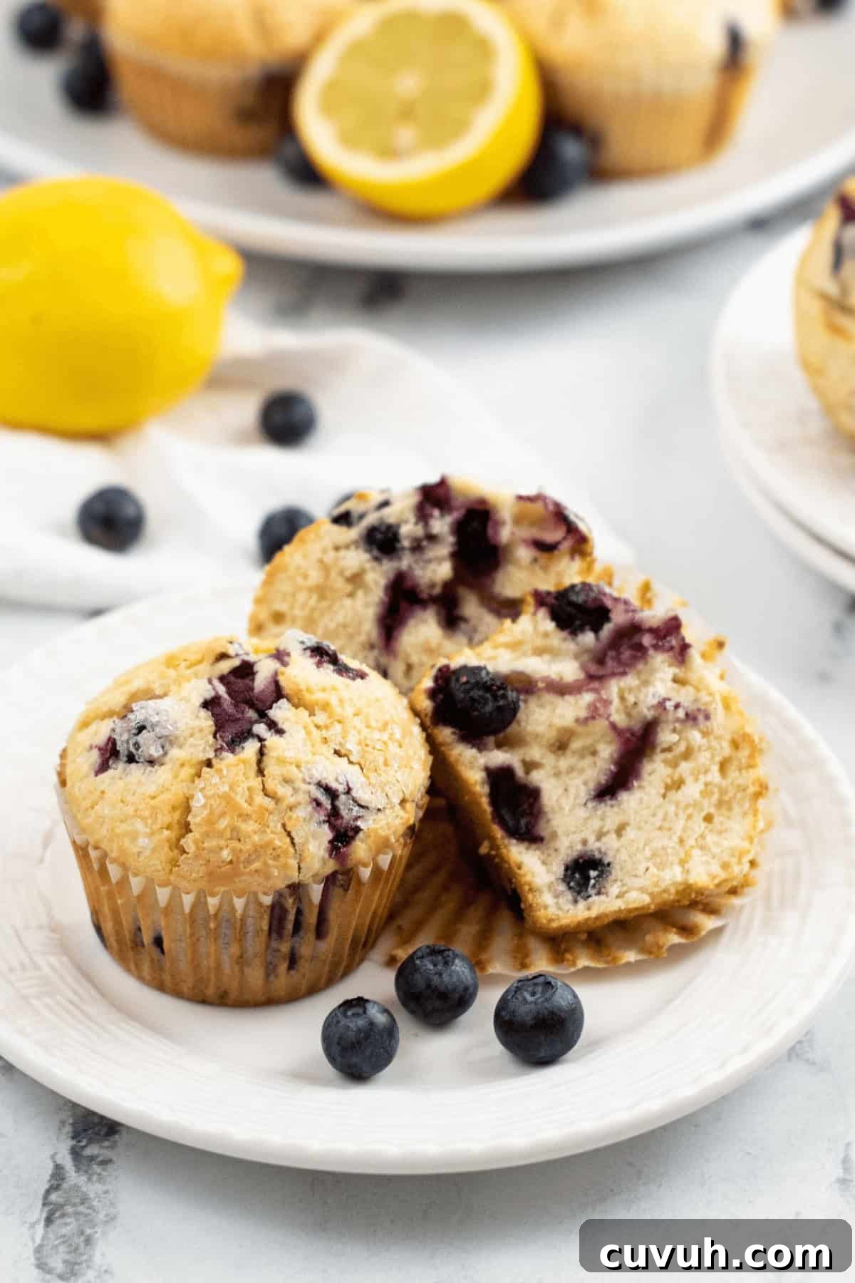 Lemon Blueberry Muffins with Luscious Citrus Glaze 7 A plate with the breakfast halved, surrounded by fresh blueberries and a lemon. Additional muffins and a lemon are visible in the background.