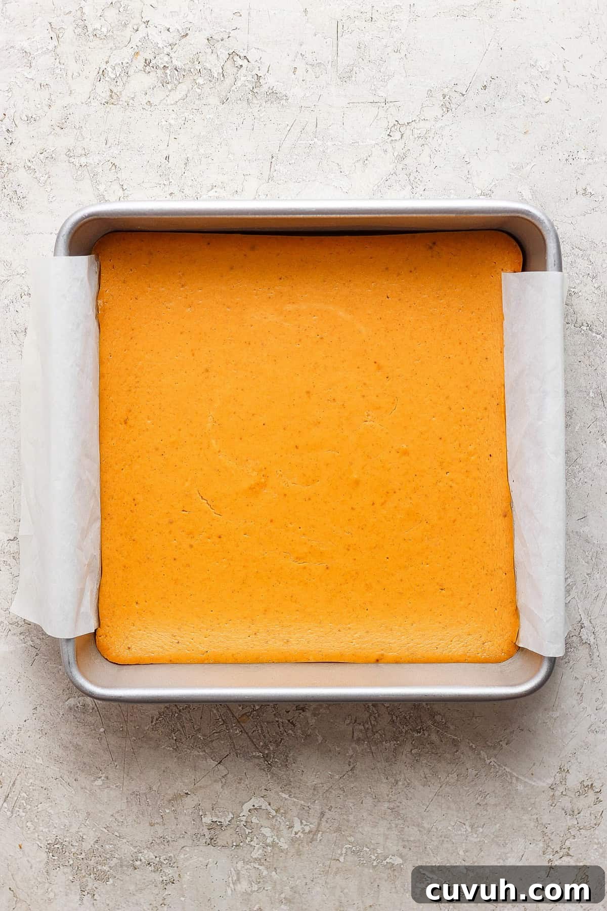 A square pan with a light golden-brown baked cake lined with parchment paper on a textured, light-colored surface.