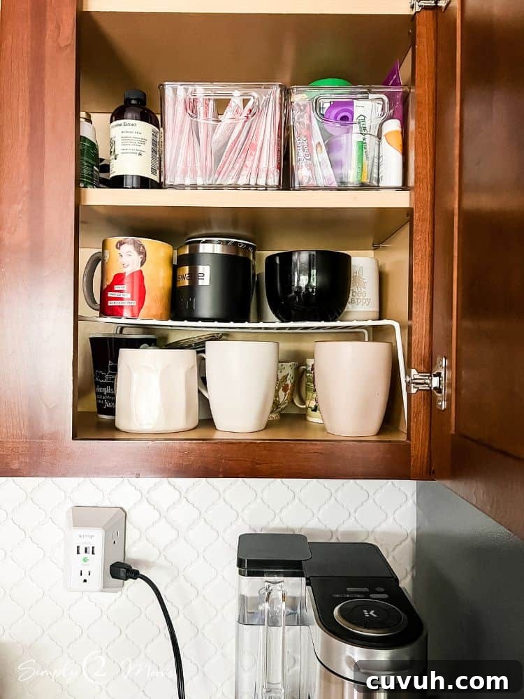 Cabinet with mugs stacked on a wire shelf insert. Clear plastic storage containers hold coffee and hot cocoa supplies on second shelf.