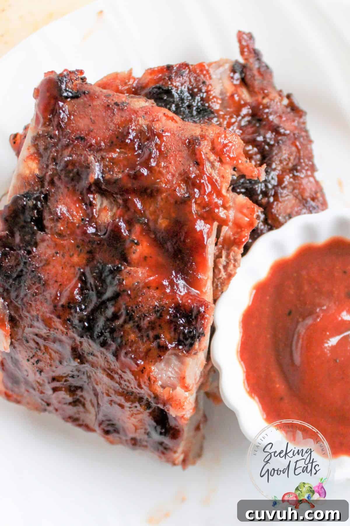 Two sections of cooked and grilled baby back ribs on a white plate with a cup of BBQ sauce.