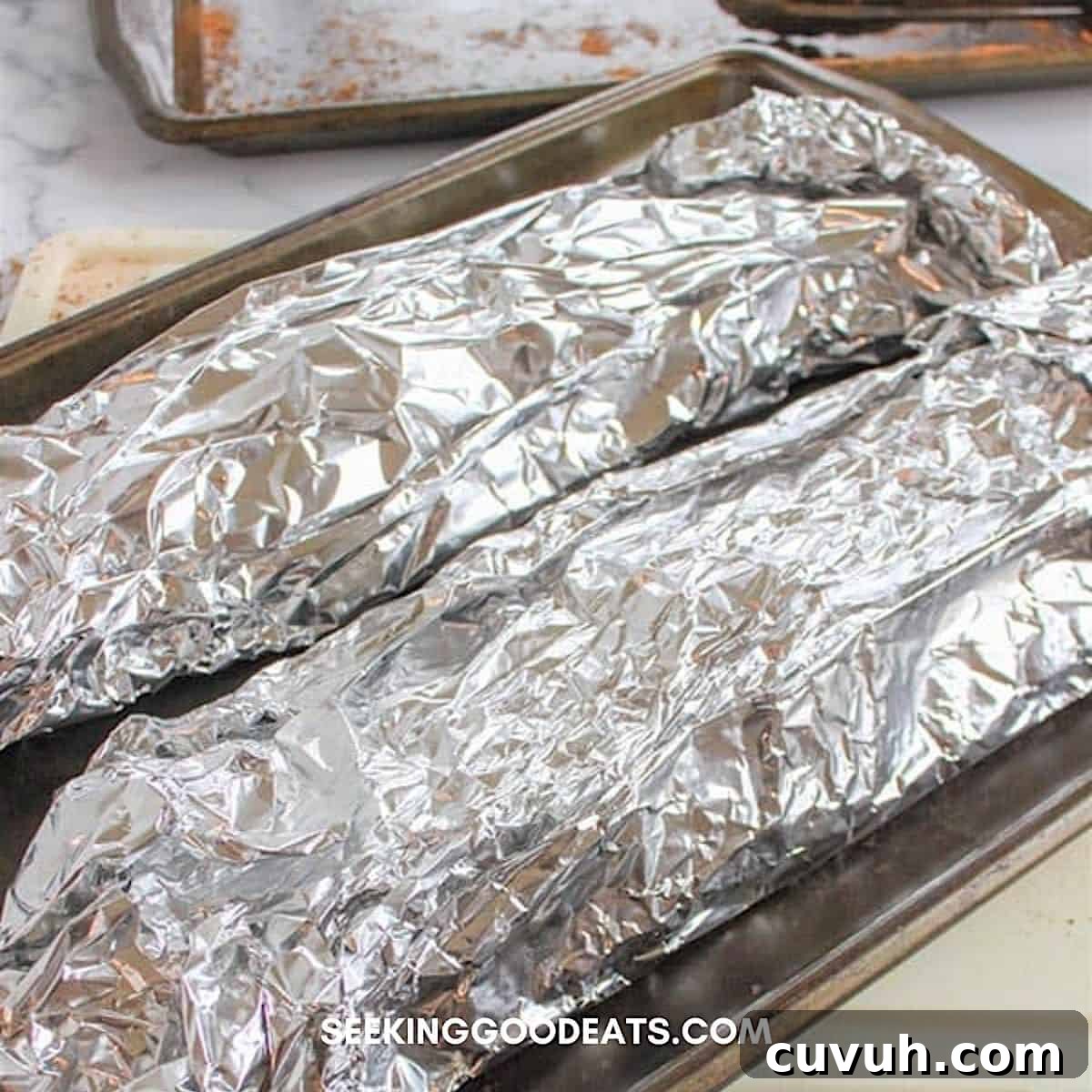 Two racks of baby back ribs tightly wrapped in foil sitting on a baking sheet.