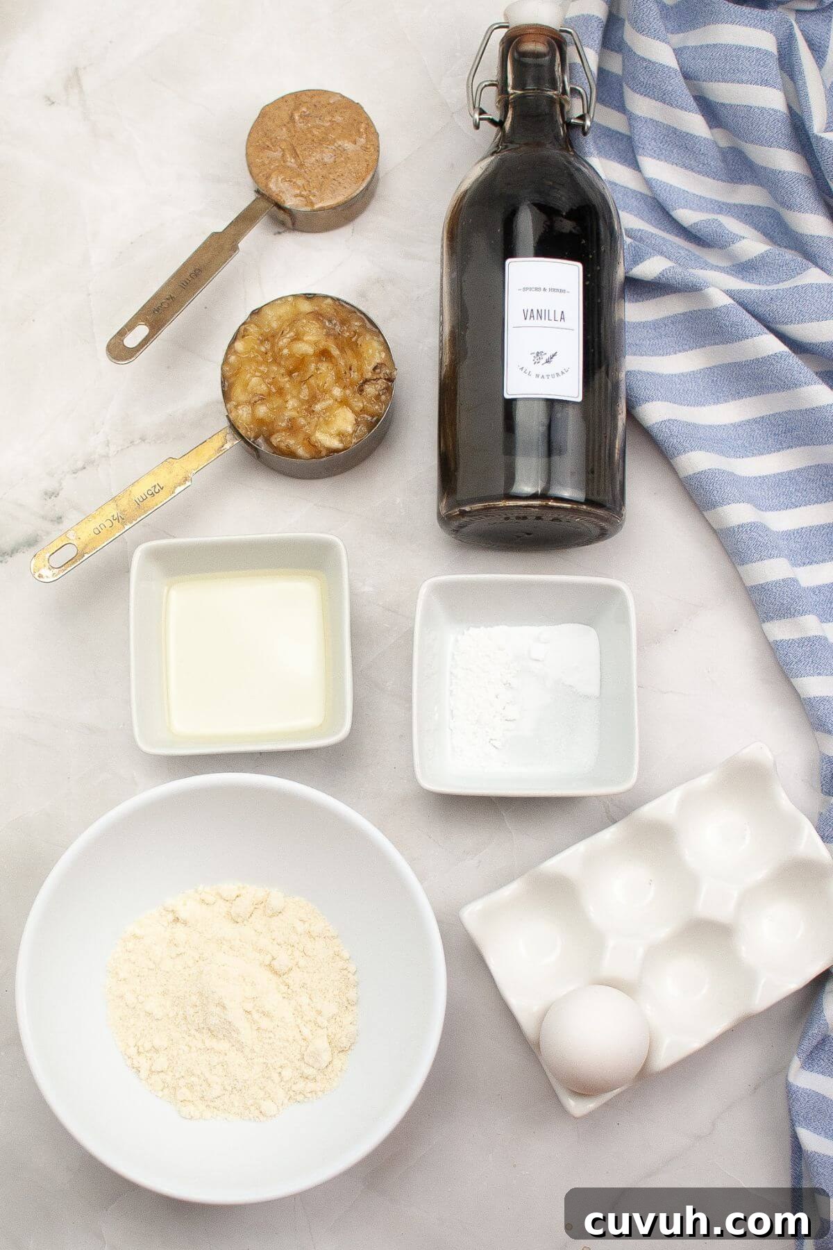 Ingredients on a countertop: vanilla extract bottle, mashed bananas, peanut butter, flour, egg, oil, baking powder, and a striped cloth.