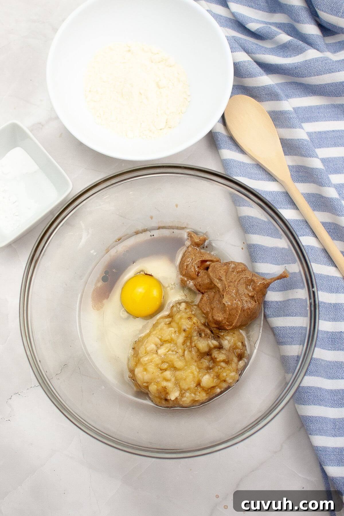 Ingredients in a glass bowl: mashed banana, egg, and peanut butter. Nearby are bowls with flour, baking soda, and a wooden spoon on a striped cloth.