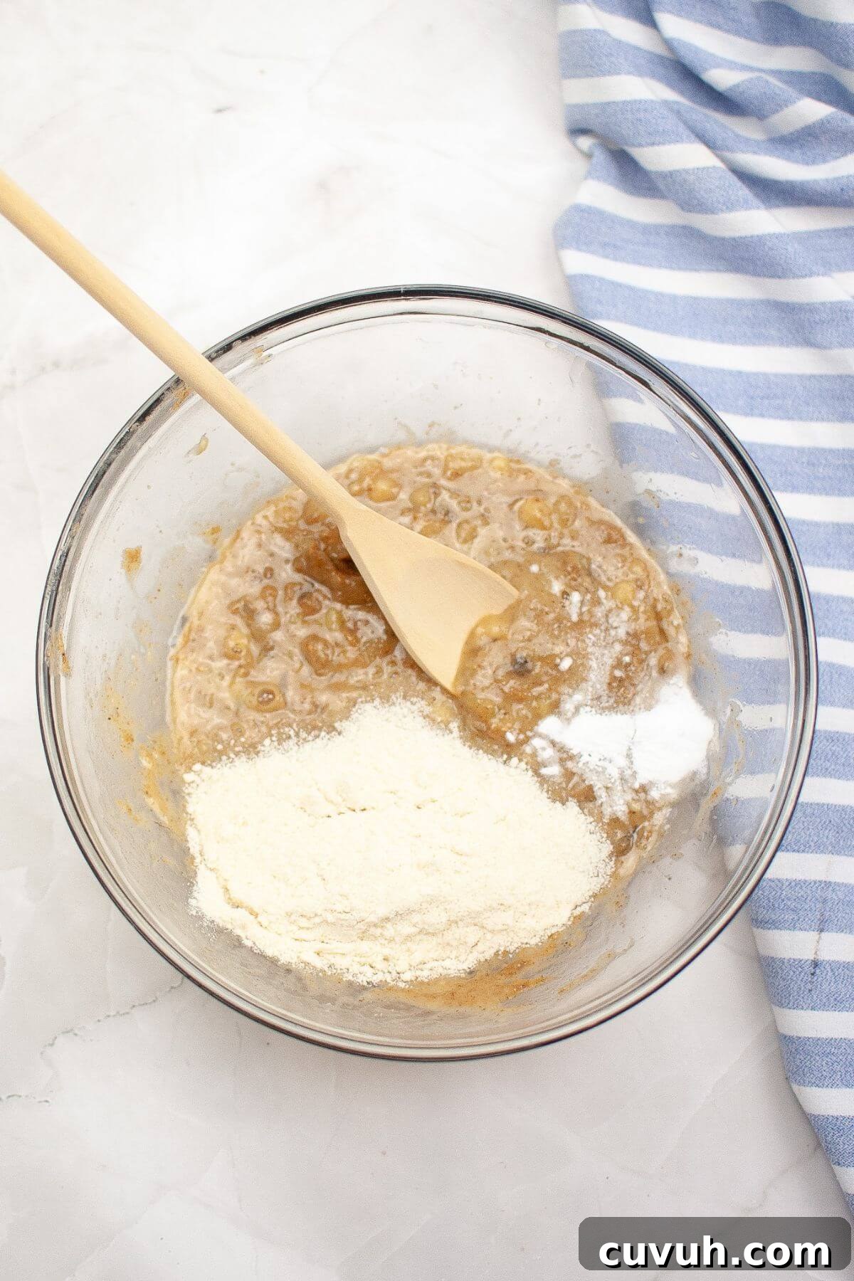 A glass bowl with batter and flour being mixed with a wooden spoon, next to a blue and white striped cloth.