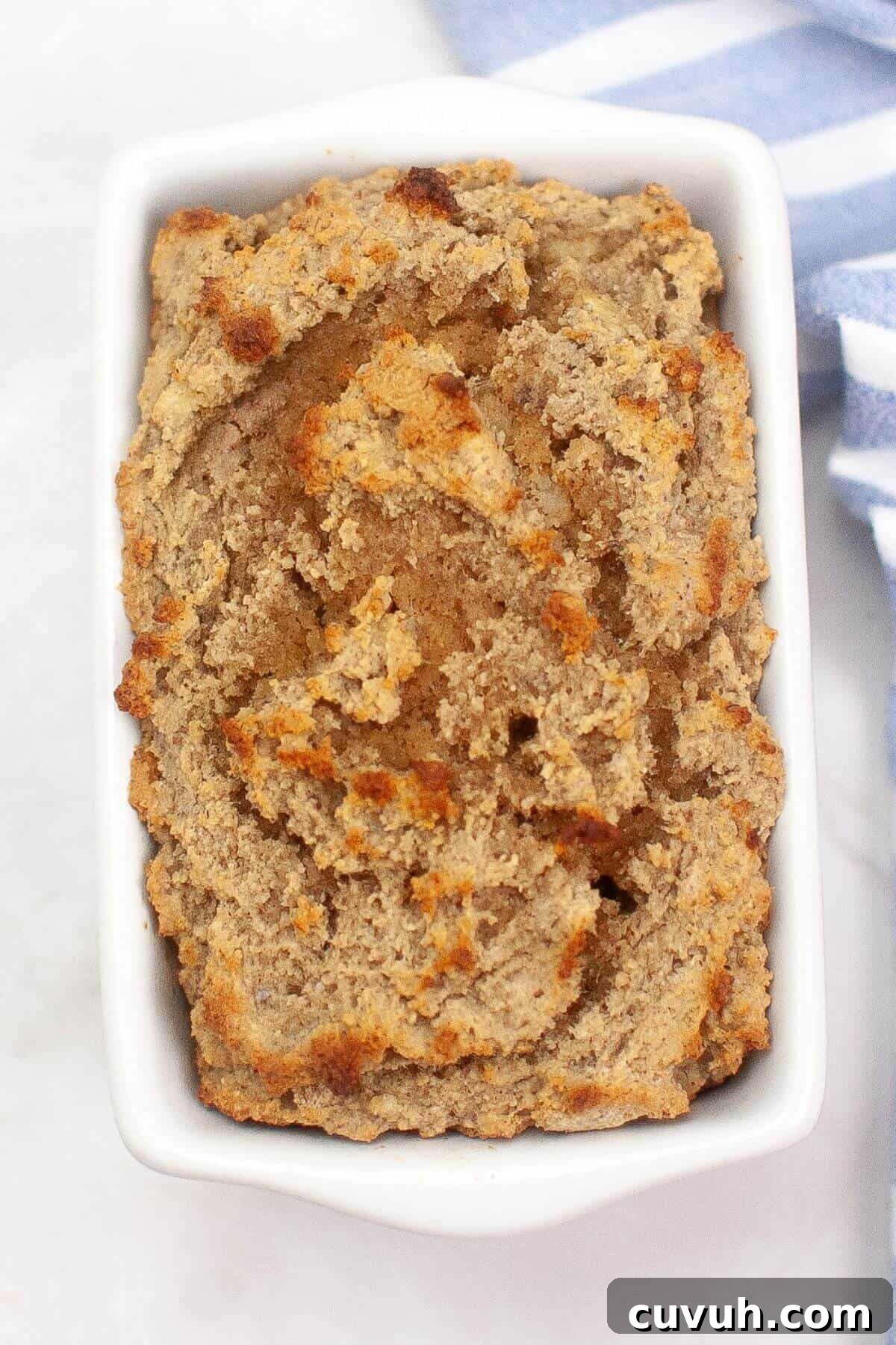 A rectangular white dish filled with freshly baked banana bread, featuring a golden-brown, textured crust. A folded striped cloth is partially visible beside it.