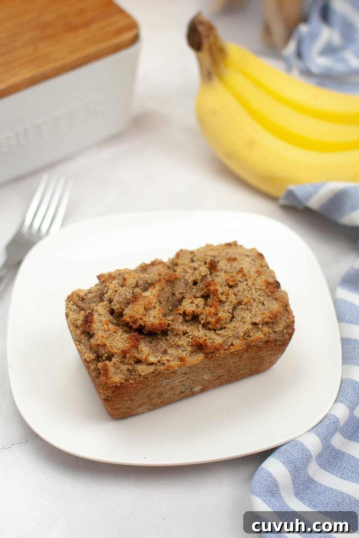 A loaf of banana bread on a white plate. A yellow banana, a striped cloth, and a fork are nearby.