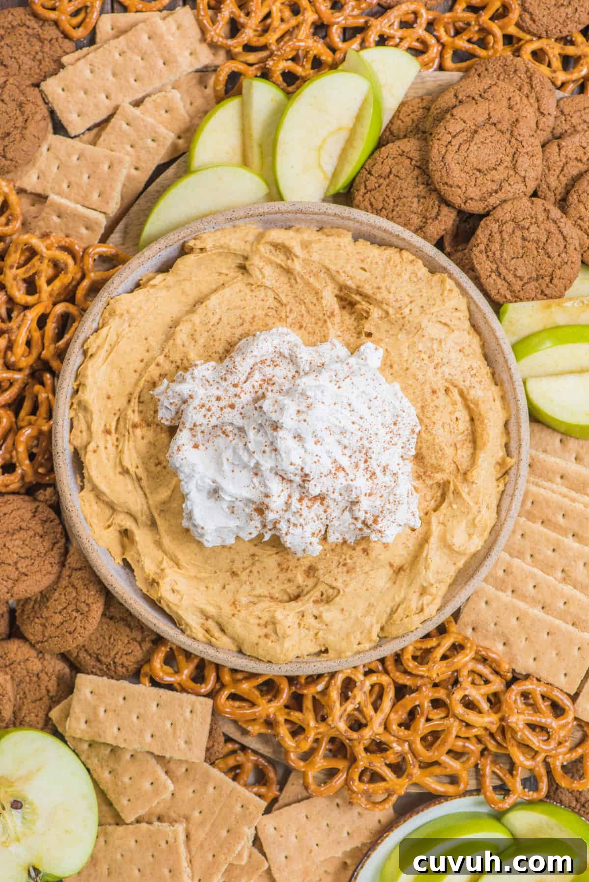 pumpkin dip with whipped cream topping in a bowl surrounded by piles of pretzels, gingersnaps, graham crackers and green apple slices