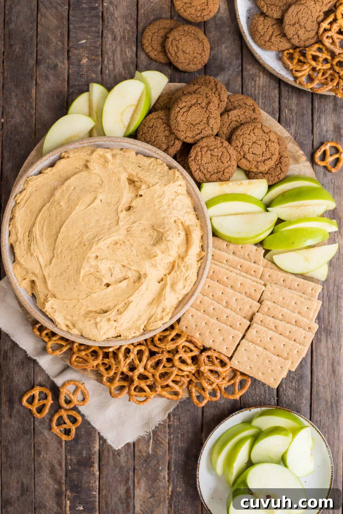 pumpkin dip in a bowl surrounded by piles of pretzels, gingersnaps, graham crackers and green apple slices on wooden background 